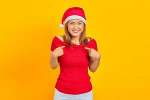 Alegre joven vistiendo gorro de navidad apuntando a sí misma sobre fondo amarillo foto