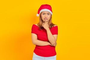 Atractiva joven asiática vistiendo gorro de navidad y haciendo gesto de silencio sobre fondo amarillo foto
