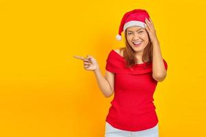 Smiling young woman wearing christmas hat with open mouth holding head in hands and pointing aside