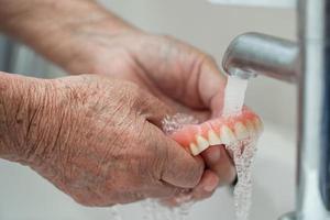 Paciente mujer mayor asiática sosteniendo y lavando la dentadura en la sala del hospital de enfermería foto