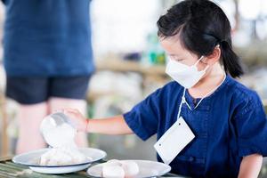Girl wearing white face mask. Child sitting in food preservation class. Kid use handle of stainless steel cup and pour ingredients together.