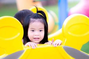 Asian child playing hide and seek on playground.  Preschool girl peek behind yellow player machine with friend or her brethren. Happy children sweet smile with playing. photo