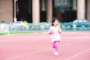Active girl running have fun on red track in stadium. Happy kid smiling and laughing. Child exercise. Baby wearing pink dress is 3-4 years old. photo
