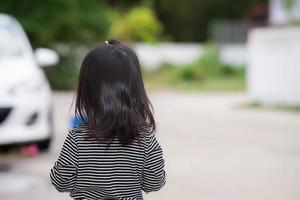 Rear view little girl stood on road. In front her there was blurry car. Concept safety child on public road. Kid wear diagonal pattern shirts with black and white stripes. photo