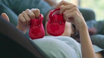 Mujer embarazada con pequeños zapatos rojos para caminar sobre el vientre mientras está acostado en el regazo del marido en la sala de estar video