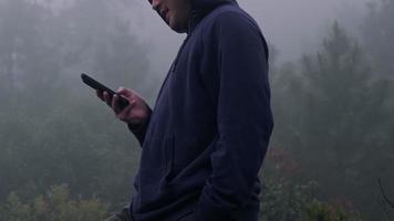 jonge man reiziger in marineblauw mannelijke hoodie sweatshirt lange mouw met behulp van een smartphone en geniet van het landschap in de bergen met dikke mist op de achtergrond. video