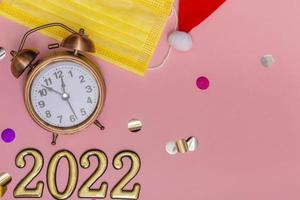 Concepto de año nuevo sobre fondo rosa reloj despertador en amarillo máscara médica números dorados 2022, cerrar foto