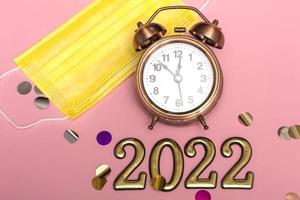 New year concept on pink background alarm clock in yellow medical mask with golden numbers 2022, close up photo