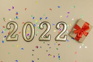 Top view on beige background of gold digits 2022 with red bow gift box decorated with multicolored sequins. Concept, layout of a New Year's card. photo