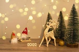 Christmas and New Year's decor. Christmas deer, numbers 2022, gnome on a sled and Christmas trees on a wooden background with Christmas lights. Christmas card