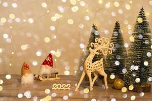 Christmas and New Year's decor. Christmas deer, numbers 2022, gnome on a sled and Christmas trees on a wooden background with Christmas lights. Christmas card photo