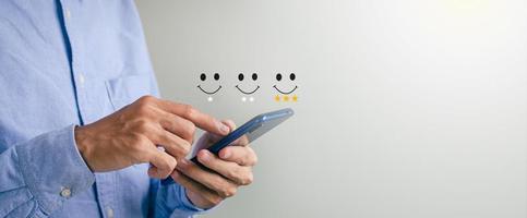 concepto de calificación de satisfacción del cliente un hombre sostiene un teléfono inteligente viendo una actuación en una pantalla de puntuación con un símbolo de oro de tres estrellas y una cara sonriente para su satisfacción. foto