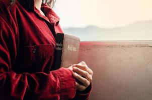 una adolescente vestida con una blusa roja está orando por la bendición de dios. tenga una vida mejor una mujer abraza la biblia con una biblia junto a la ventana, hermosa vista, una crisis en la vida cristiana