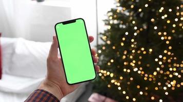 homem segurando um smartphone com tela verde chromakey sobre a árvore de Natal com luzes