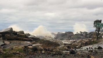 olas rompiendo contra las rocas en el borde del lago Michigan. cielo nublado y con volutas de azul. acera cubierta de escombros rocosos. agua turbulenta salpicando alto en las rocas