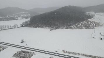 Aerial view of snow covered farm field in valley in Wisconsin. Forest covering mountainside. Trails on side of hill. Highway seen in foreground with one directional sign.