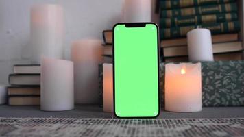 Modern smartphone standing on surface with green screen chromakey with candles and books at the background video
