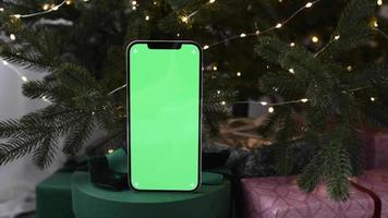 Modern smartphone standing on gifts with green screen chromakey near Christmas tree lights on background video