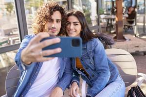 Pareja árabe tomando fotos selfie con su teléfono inteligente, sentada en la terraza de un bar.