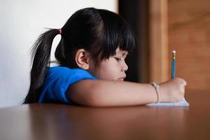A cute little Asian girl is writing a book with a pencil on the table. photo