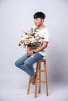 A young man in a white t-shirt sits on a high chair and holds flowers. photo