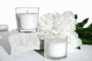 white scented soy wax candles on a natural stone and peony flowers. floral fragrance for cozy home. aromatherapy and wellness concept photo