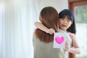 Asian mother hug her cute daughter that give handmade greeting card with colourful heart symbol to surprising her at home photo