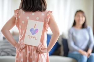 linda chica asiática esconde una tarjeta de felicitación hecha a mano con la palabra amo a mamá para sorprender a su madre en casa