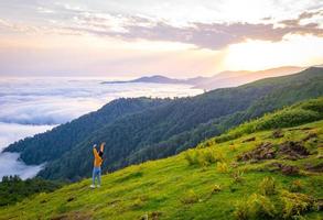 Persona femenina de alegría mira a la cámara con las manos extendidas y el paisaje escénico de la montaña gomismta por encima de las nubes con el amanecer en backgorun