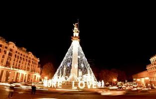 Cars pass on roundabout leaving light trails with decoration lights on statue in freedom square while tourist taking photographs photo