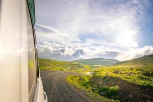 White car drives on caucasian green wilderness with sky and meadow view. Copy paste road trip background photo