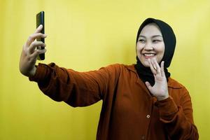 Beautiful young asian muslim woman with hands holding smartphone, calling someone, making video call, taking selfie, smiling confident, enthusiastic and cheerful isolated, advertising concept photo