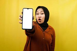 Beautiful young asian muslim woman shocked, surprised, wow expression, hand holding smartphone with white or blank screen, promoting app, promoting product, presenting something, isolated photo