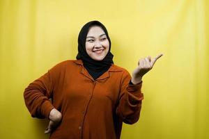 Beautiful and cheerful young asian muslim woman, with hand pointing at empty space, presenting something, isolated photo