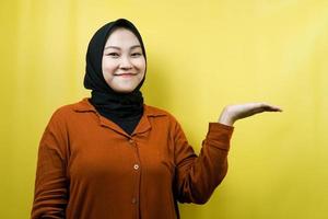 Beautiful young asian muslim woman with hands showing and presenting something in the direction of empty space, smiling confidently, excitedly, facing the camera, isolated, advertising concept photo
