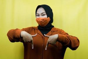 Muslim woman wearing mask, hand pointing down, presenting something, click the link below, isolated photo