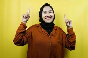 Beautiful young asian muslim woman with hands pointing up presenting something, looking at camera isolated, advertising concept photo