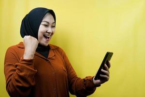 Beautiful young asian muslim woman with hands holding smartphone, smiling confident, enthusiastic and cheerful, hands clenching, punching, victory, success, good work, isolated, advertising concept photo