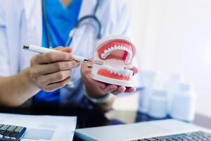 Dentista concentrado sentado a la mesa con muestras de mandíbula modelo de diente y trabajando con tableta y portátil en el consultorio dental foto