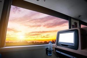 dentro del autobús que tiene un asiento trasero en blanco con pantalla lcd para entretenimiento con una botella de agua y vista de la ventana del hermoso paisaje, la naturaleza con el cielo, la nube, el atardecer, el concepto de viaje por carretera de turismo de figura. foto