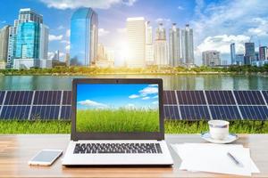 Mockup image of laptop with Alternative green clean energy concept on screen and smartphone,coffee cup on wooden table of solar panel  with City and skyscrapers background.