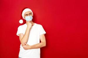 serious man in thoughtful pose holds his hand under his chin in Christmas Santa Claus hat isolated on red background with space for text concept - brainstorming, gift selection, holiday ideas, holiday photo