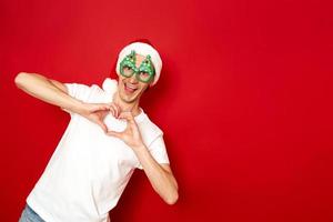 Lovely smiling crazy man shows heart gesture over body expresses love to someone romantic feelings, wears Christmas hat on head white t-shirt. concept - New Year, body language, people. space for text photo