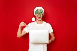 an excited smiling man in Santa Claus Christmas hat points with his index finger laptop in his hands recommends online purchases, sales, promotions, discounts isolated on red background space for text photo