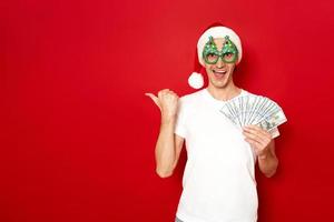 excited, cheerful, surprised man in Santa New Year hat with wad of dollars money points finger at free empty area isolated on red background space for text concept - credit, cashback, bank, investment photo