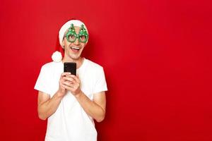 Concept - people Smiling excited young man in casual blank white t-shirt xmas hat christmas tree glasses posing on red studio wall. Using mobile cell phone typing sms message browsing looking aside up photo