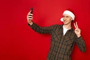 Man takes selfie makes video call points at camera of smartphone enjoys spare time dressed in casual clothes and christmas hat shows peaceful gesture isolated on red background concept - communication photo
