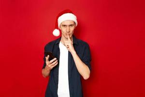 Shh. Keep secret. No sound mute silent confidential, privacy quiet, shush hissing hush concept. serious with forefinger by his lips, phone in hands. Festive holly jolly xmas. isolated studio red wall photo