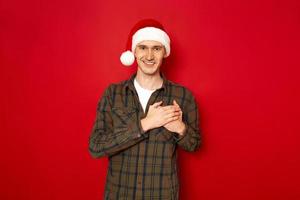 Indoor shot of handsome man keeps hands on chest, being cordial and friendly, expresses gratitude and thankfulness, has pleasant smile, dressed in casual outfit xmas hat, isolated on red studio wall photo