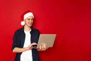 Laughing young man in casual blue shirt christmas hat posing isolated on red background studio portrait. People sincere emotions lifestyle concept. Mock up copy space. Working on laptop pc computer photo
